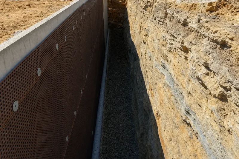 A brown dimpled drainage sheet fixed over black tanking on a below-ground basement wall, slotted agricultural pipe in washed gravel at the footing, a layered sandstone cut face across the open trench — illustrative example
